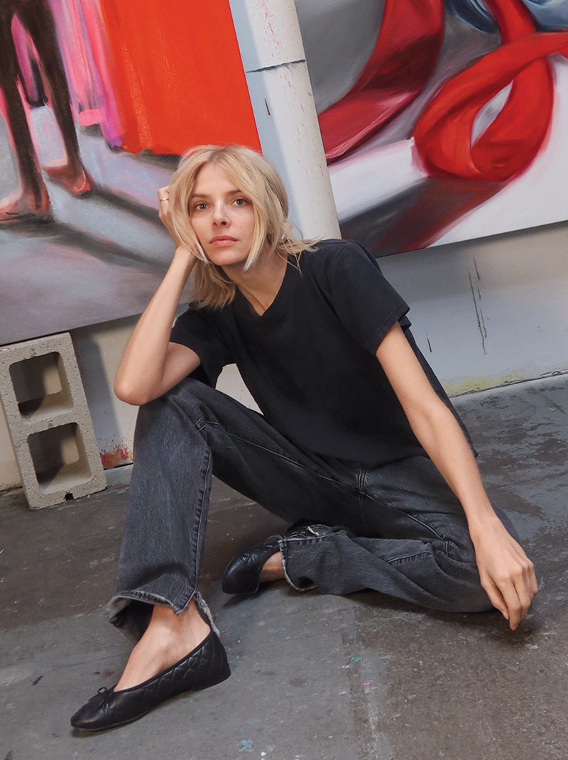 Artist Amanda Wall poses on the floor of her studio wearing light gray pants and a black t-shirt.