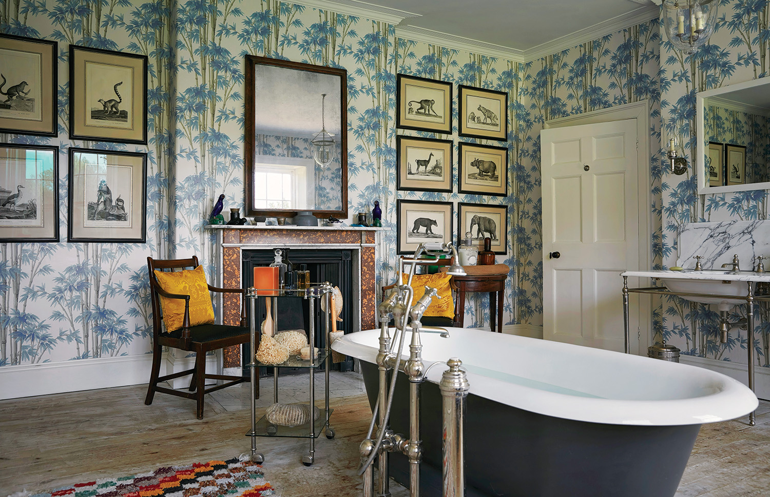 In the primary bath reimagined by Ben Pentreath, clad in a Zuber wallpaper, antique animal prints flank a 19th-century burr-walnut mirror above the fireplace. The pendant lantern is by Hector Finch, while a Charles Edwards sconce is mounted next to a bespoke mirror over the vanity, which is by Drummonds.