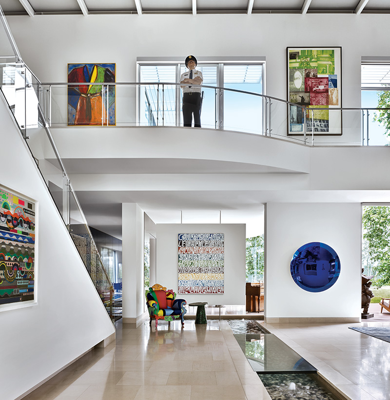 A work by Beatriz Milhazes is mounted along the stairs near the entry of a home designed by Ernest de la Torre, where at the far end a Mel Bochner text painting overlooks an Alessandro Mendini chair and a malachite table. A cobalt-hued Anish Kapoor wall sculpture shimmers across the water feature. On the mezzanine above, a Marc Sijan sculpture of a security guard stands sentry between a Jim Dine “Robe” painting and a print by Robert Rauschenberg.