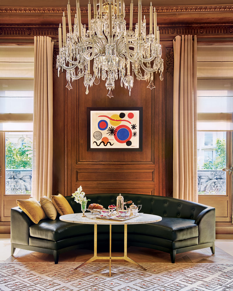 An Alexander Calder painting, displayed between windows curtained in a Dedar fabric, adds a colorful note to the dark-paneled living room, where an 18th-century La Granja chandelier dazzles above the Hazarian-designed leather sofa, which curls around a marble-and- bronze table topped with Hermès coffee cups.