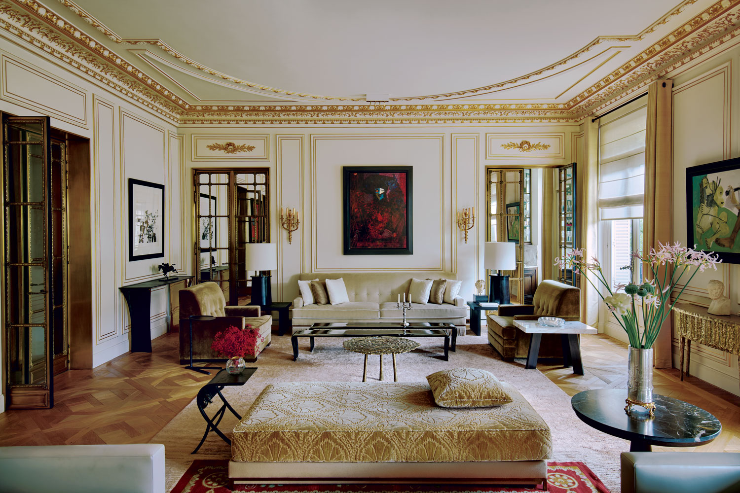 In the second living room, ornamented with gilt paneling and moldings by Atelier Mériguet-Carrère, a Max Ernst painting flanked by 19th-century sconces overlooks a seating area where a sofa, chairs, and multiple tables by Hazarian are arrayed around long cocktail tables by Eric Schmitt, joined by a pair of Liaigre lamps. At the room’s center, a custom daybed is covered in a Fadini-Borghi fabric, while on the sides, artworks by Zao Wou-Ki (left) and Pablo Picasso are mounted above consoles by Hazarian.