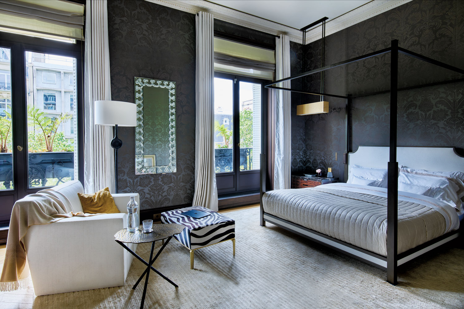 The primary bedroom features a wall covering and curtain fabric by Dedar, bed linens by Frette, a mirror by Lalique, and a rug by JD Staron. Hazarian designed the lounge chair, lamp, side table, and hide-upholstered ottoman.