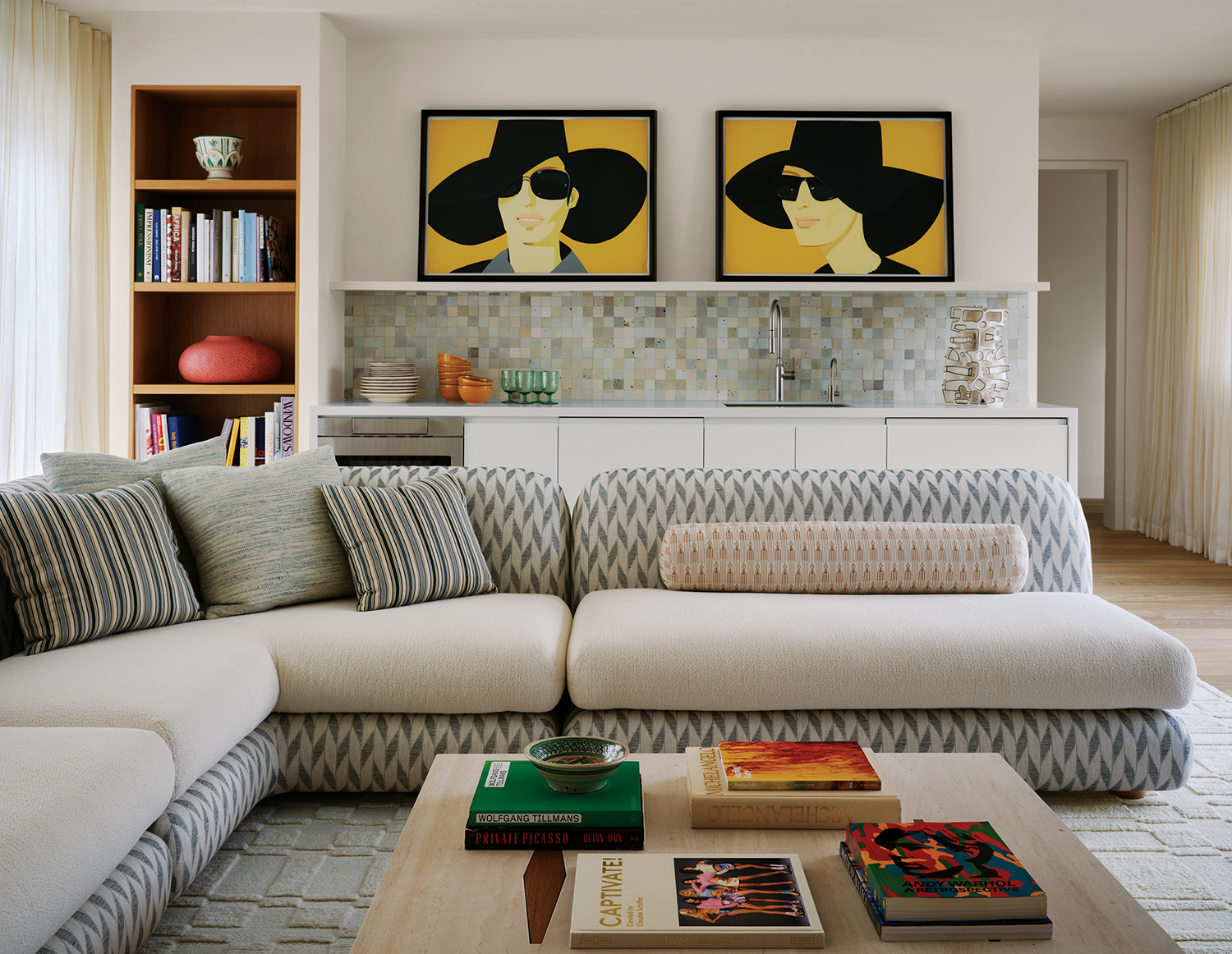 In the upstairs family room, a custom sofa clad in Rosemary Hallgarten and Rose Tarlow Melrose House fabrics and accented with pillows made of John Rosselli & Associates textiles wraps around a Jorge Zalszupin by Etel table from the Invisible Collection. Alex Katz prints animate the bar area, which features a backsplash of Clé tiles.