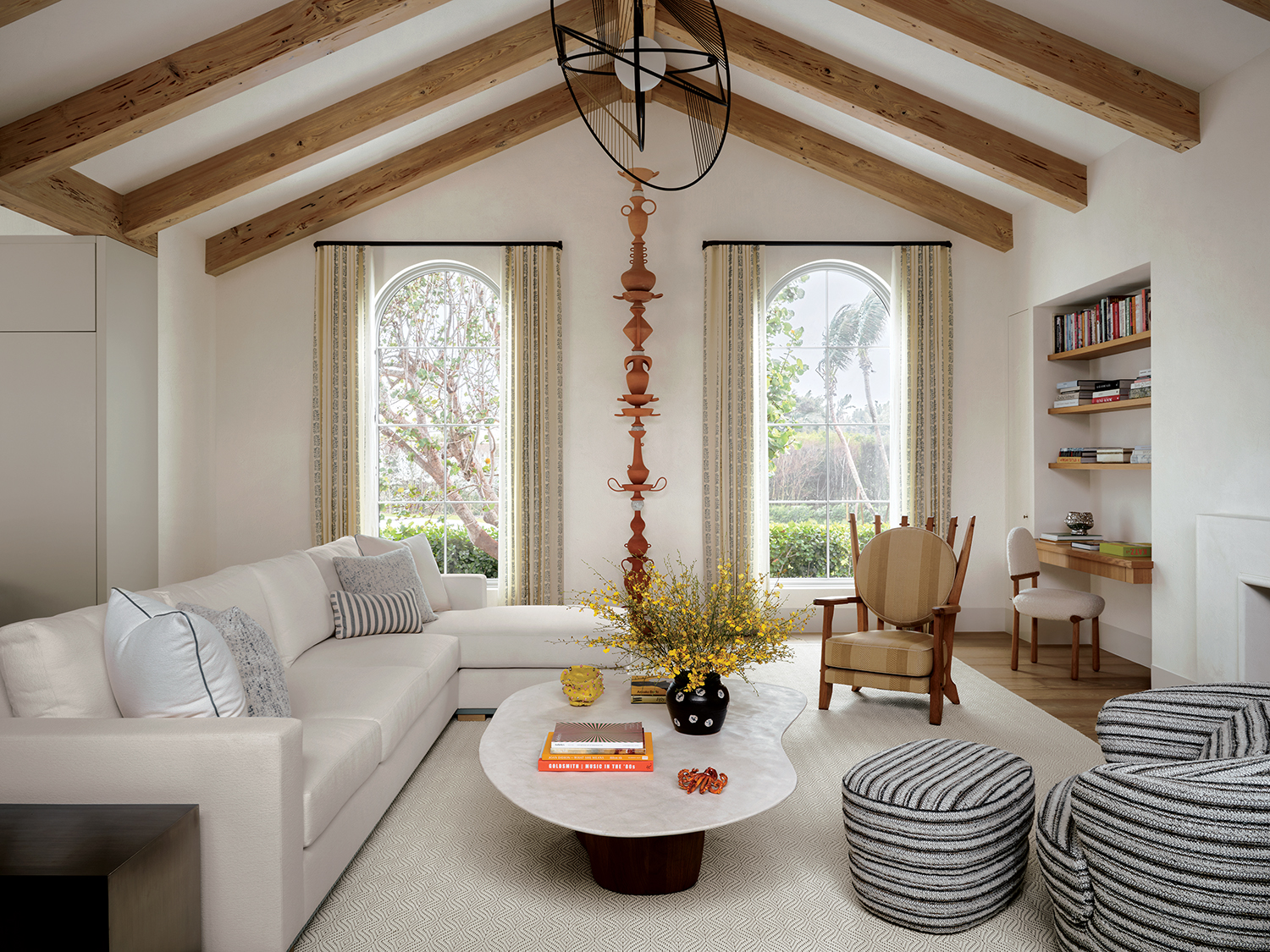 A towering Cammie Staros sculpture stands between windows curtained in a Rose Tarlow Melrose House fabric in the family room, which is crowned by a Jean-Louis Deniot light fixture. The custom sofa and cocktail table are joined by a lounge chair and an ottoman by Bunn Studio for Radnor in a Toyine Sellers stripe and a Guillerme et Chambron armchair; the desk chair is by Hubert Le Gall from Twenty First Gallery.