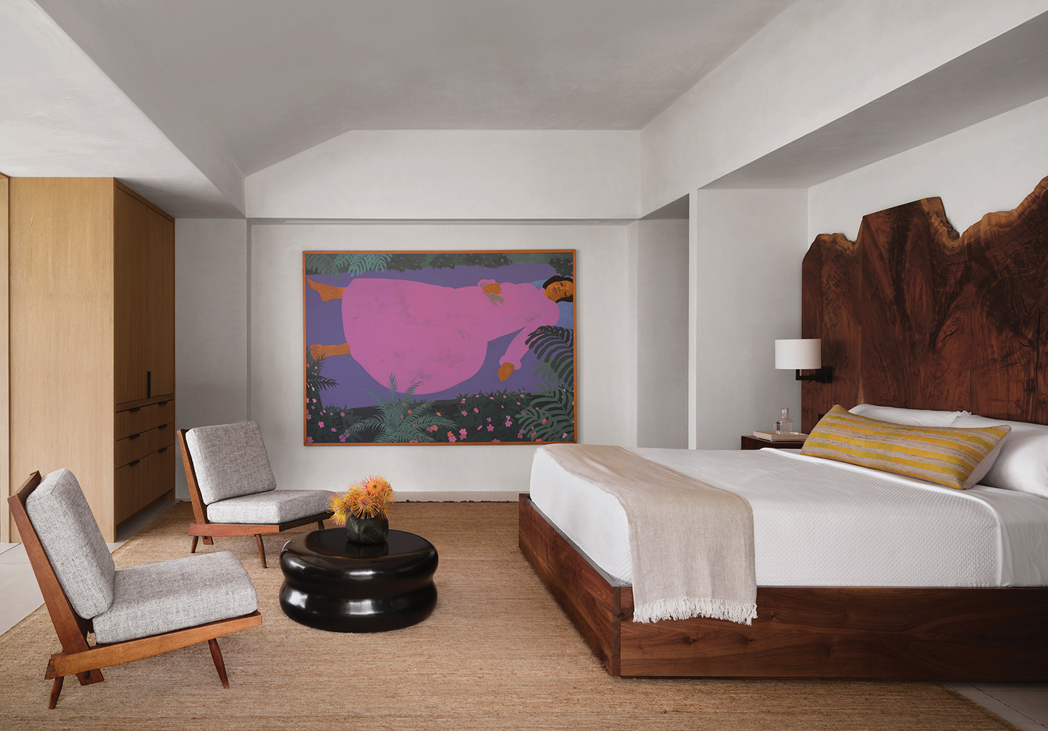 A Pegge Hopper painting enlivens the primary bedroom, where Hollis designed the teak dresser cabinet and the walnut bed featuring integrated nightstands and a monumental slab headboard with a pair of Liaigre sconces; 1960s George Nakashima lounge chairs join a Liaigre table.