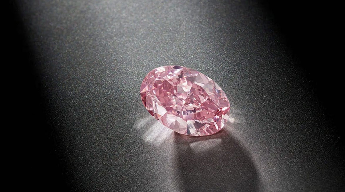 Oval pink diamond with intricate facets placed on a dark textured surface, reflecting light.